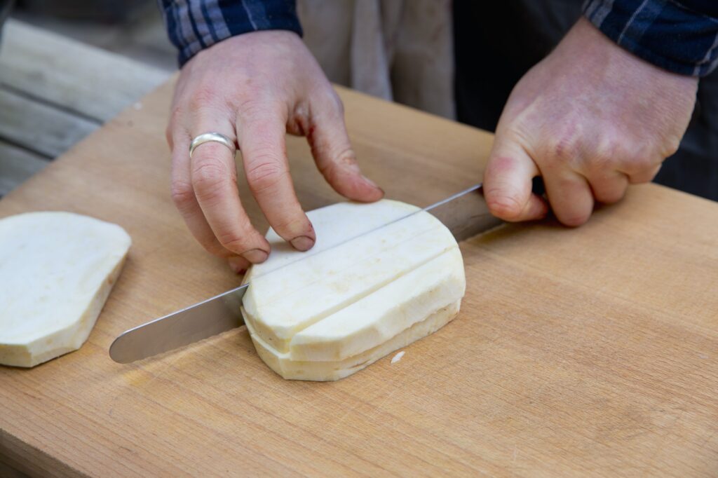A man with a long knife chopping celeriac into cubes on a chopping board.